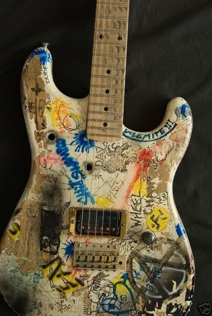 A punk guitar. White guitar body, beat up, with black handwritten anarchy symbol and various handwriting